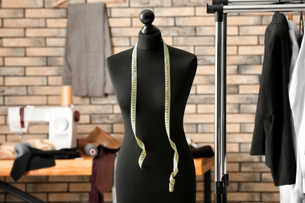 Mannequin with measuring tape in front of clothes rack, showcasing fashion design and tailoring concept.